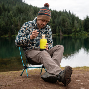Man sitting nearby an alpine lake eating ramen out of EcoSimmer flameless camping stove